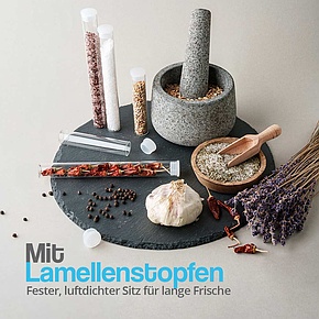 Glasröhrchen mit Lamellenstopfen, gefüllt mit Salz, Pfeffer und getrockneten Chilis, auf einer Schieferplatte mit Kräutern